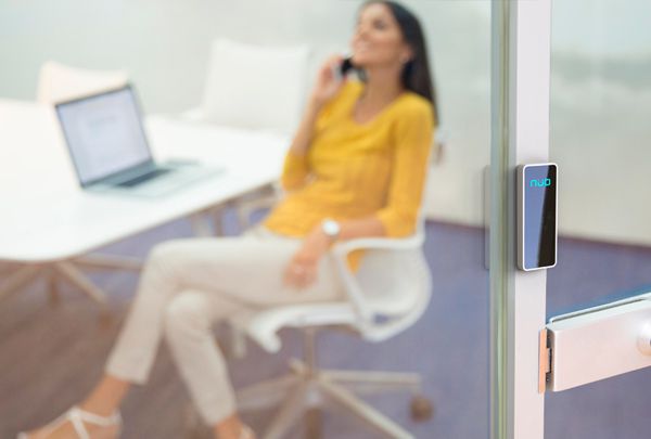 Mujer con camisa amarilla y pantalón blanco, sentada en una silla blanca, hablando por teléfono y en la puerta de entrada, hay un lector NÜO surf, lector de control de acceso de la marca NÜO